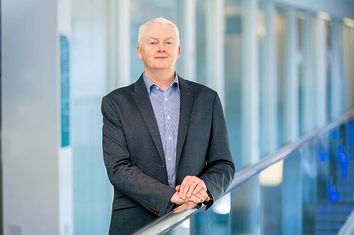 Professor Derek McGhee in formal business attire leaning against a glass baulastrade