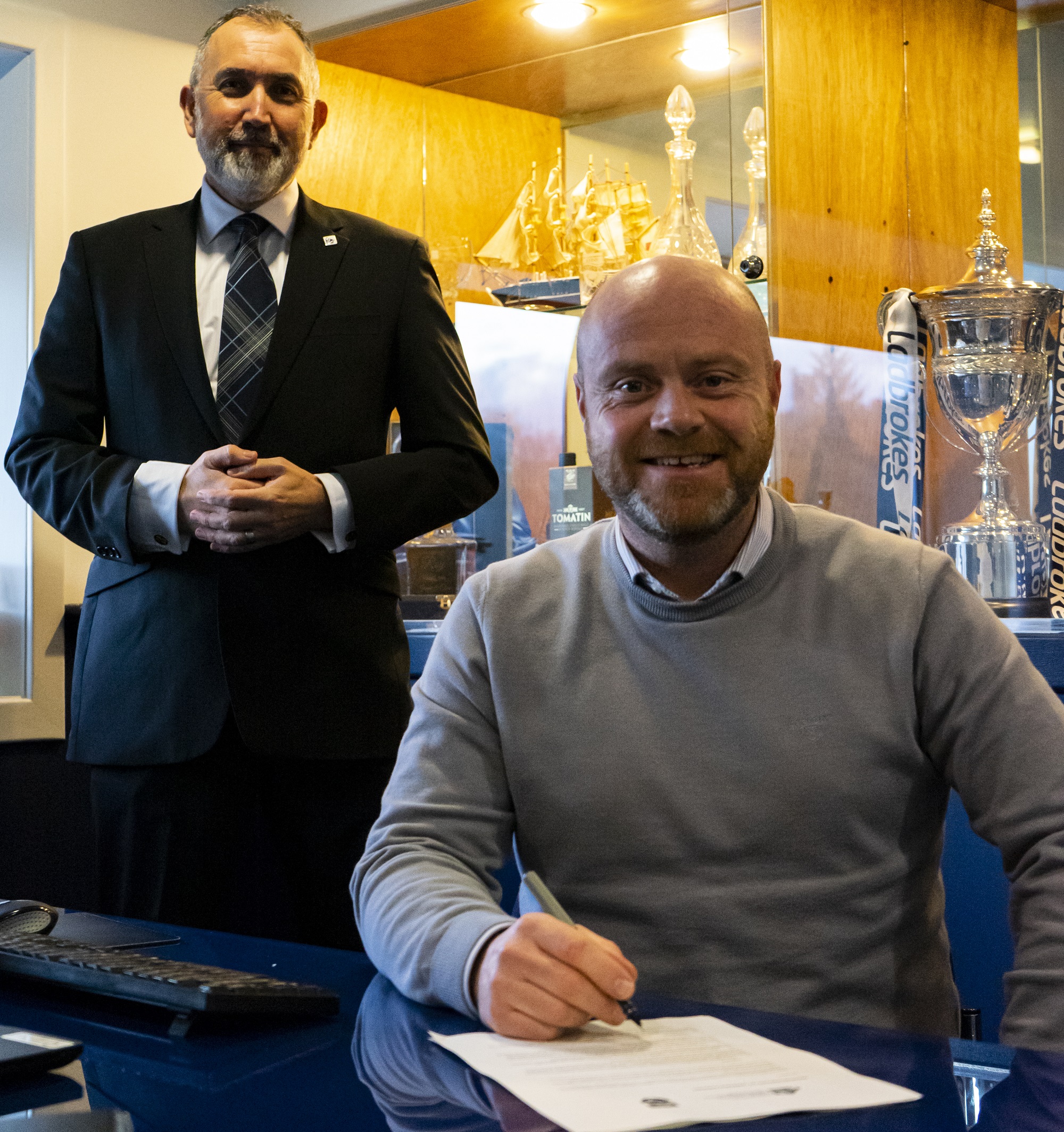 One person sitting with a pen signing a piece of paper. One person standing behind One person sitting with a pen signing a piece of paper. One person standing behind