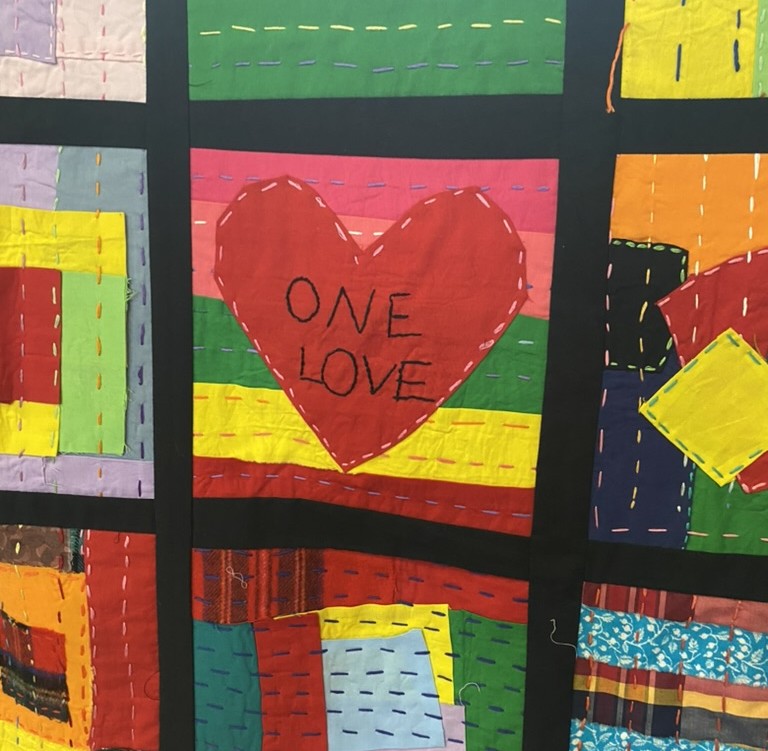 A colourful textile made up of square patches, with one prominent red heart in the centre patch surrounded by various bright colours like yellow, green, and pink.