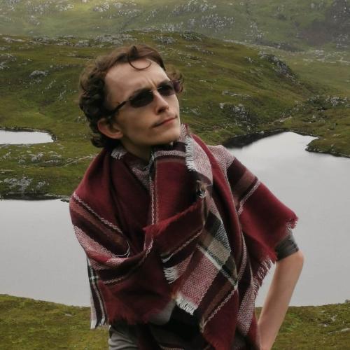 Man wearing sunglasses and scarf standing in the forefront while the background shows a green landscape and a loch Man wearing sunglasses and scarf standing in the forefront while the background shows a green landscape and a loch